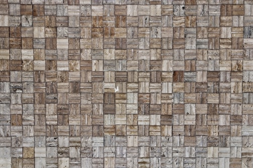 A close-up view of a textured wall composed of small rectangular stone tiles arranged in a grid pattern. The colors of the tiles vary, featuring different shades of brown, beige, and gray. The surface shows natural stone patterns with slight variations in texture and appearance.