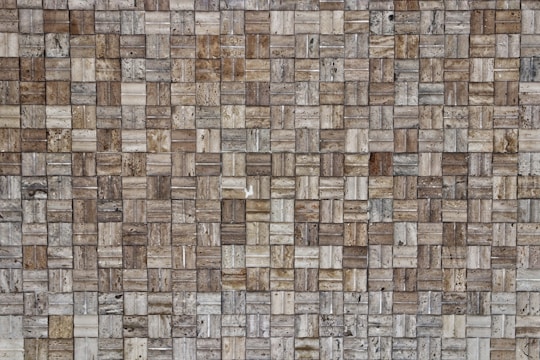 A close-up view of a textured wall composed of small rectangular stone tiles arranged in a grid pattern. The colors of the tiles vary, featuring different shades of brown, beige, and gray. The surface shows natural stone patterns with slight variations in texture and appearance.