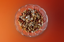 A clear glass bowl filled with an assortment of mixed nuts and seeds, including almonds, peanuts, and pumpkin seeds. The background is a gradient of warm orange and red tones.