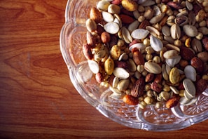 A clear glass bowl filled with a variety of mixed nuts including almonds, pistachios, and chickpeas on a wooden surface.