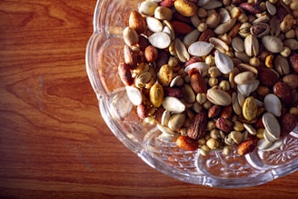 A clear glass bowl filled with a variety of mixed nuts including almonds, pistachios, and chickpeas on a wooden surface.