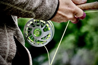 A close-up view of a person holding a fishing rod with a focus on the reel. The hand is wearing a woolen glove, suggesting cool weather. The reel is metallic with green accents, and there is greenery in the blurred background, implying an outdoor setting.