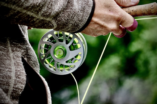 A close-up view of a person holding a fishing rod with a focus on the reel. The hand is wearing a woolen glove, suggesting cool weather. The reel is metallic with green accents, and there is greenery in the blurred background, implying an outdoor setting.