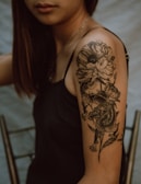 A close-up view of a woman's upper arm featuring a detailed tattoo. The tattoo depicts a large flower with petals and leaves intertwined with a fierce dragon. The woman is wearing a black sleeveless top, and her hair is straight and brown. The background is blurred with neutral colors, bringing focus to the intricate tattoo design.