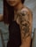 A close-up view of a woman's upper arm featuring a detailed tattoo. The tattoo depicts a large flower with petals and leaves intertwined with a fierce dragon. The woman is wearing a black sleeveless top, and her hair is straight and brown. The background is blurred with neutral colors, bringing focus to the intricate tattoo design.