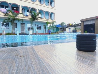 A poolside scene with a modern building in the background, featuring several circular balconies. There are palm trees and a clear blue sky, creating a tropical atmosphere. Additionally, a small black speaker is placed on the wooden deck near the pool.