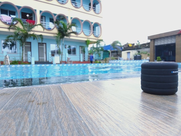 A poolside scene with a modern building in the background, featuring several circular balconies. There are palm trees and a clear blue sky, creating a tropical atmosphere. Additionally, a small black speaker is placed on the wooden deck near the pool.