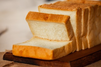 A loaf of sliced white bread rests on a wooden cutting board. The bread has a soft, fluffy texture with a golden-brown crust. The board is set on a burlap cloth, adding a rustic feel.