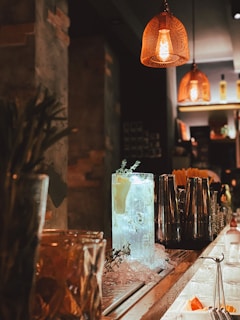 Dimly lit bar scene with a focus on a cocktail glass filled with ice, garnished with lemon and herbs, set on a counter surrounded by metal shakers and various bar tools. Warm, inviting orange light from overhead lamps casts a cozy glow.