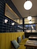 A cozy restaurant interior features a long mustard yellow upholstered bench with geometric patterned cushions. The wall is covered with glossy dark blue tiles, and a framed sign indicates the restaurant name. Above, spherical pendant lights provide illumination, while the background showcases a bar area with hanging glassware and a decorative wall with floral patterns.