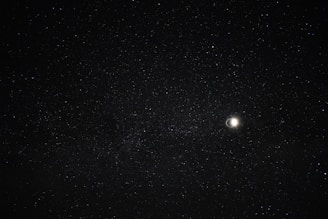 A night sky filled with countless stars, featuring a bright crescent moon to the right. The stars are spread across the dark expanse, creating a serene and vast celestial scene.