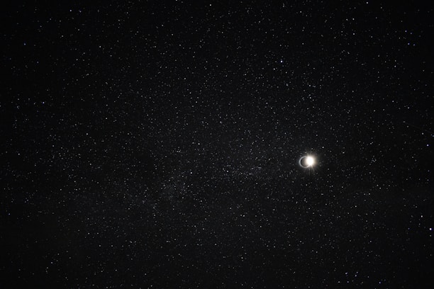 A night sky filled with countless stars, featuring a bright crescent moon to the right. The stars are spread across the dark expanse, creating a serene and vast celestial scene.