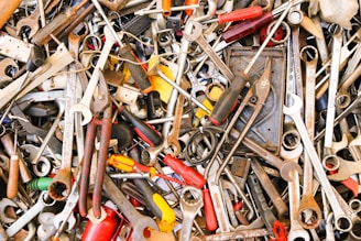 A chaotic assortment of various hand tools including wrenches, pliers, screwdrivers, and other metallic instruments. The tools are overlapping and intermingled in a haphazard pile, displaying signs of wear with rust and dust.