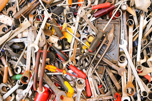 A chaotic assortment of various hand tools including wrenches, pliers, screwdrivers, and other metallic instruments. The tools are overlapping and intermingled in a haphazard pile, displaying signs of wear with rust and dust.