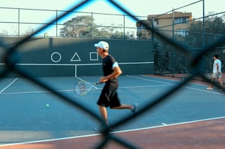 A tennis court is visible through a chain-link fence, with two people on the court. One person is in motion, holding a tennis racket, while the other is walking. Geometric shapes are painted on the wall in the background.