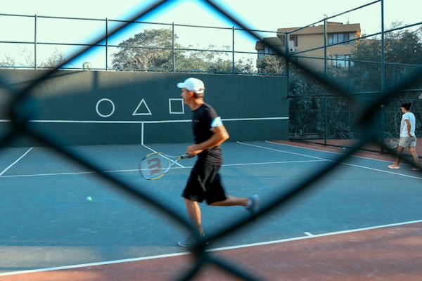 A tennis court is visible through a chain-link fence, with two people on the court. One person is in motion, holding a tennis racket, while the other is walking. Geometric shapes are painted on the wall in the background.