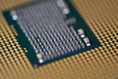 A close-up of a computer processor with numerous small metallic components and gold contact pins on a green substrate. The focus is on the central section with detailed circuitry, surrounded by a grid pattern of contacts.
