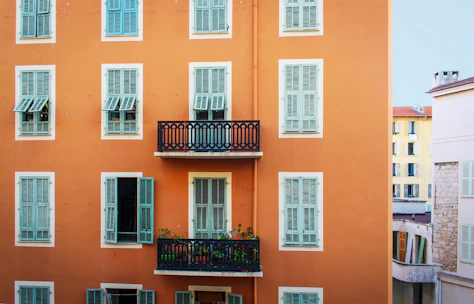 A facade of a building with an orange exterior is prominently featured, showcasing multiple windows with blue shutters. In the center, there is a small balcony with a decorative black railing and several plants. Adjacent buildings can be seen, including one with a stone wall and another with a yellow exterior.
