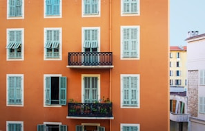 A facade of a building with an orange exterior is prominently featured, showcasing multiple windows with blue shutters. In the center, there is a small balcony with a decorative black railing and several plants. Adjacent buildings can be seen, including one with a stone wall and another with a yellow exterior.