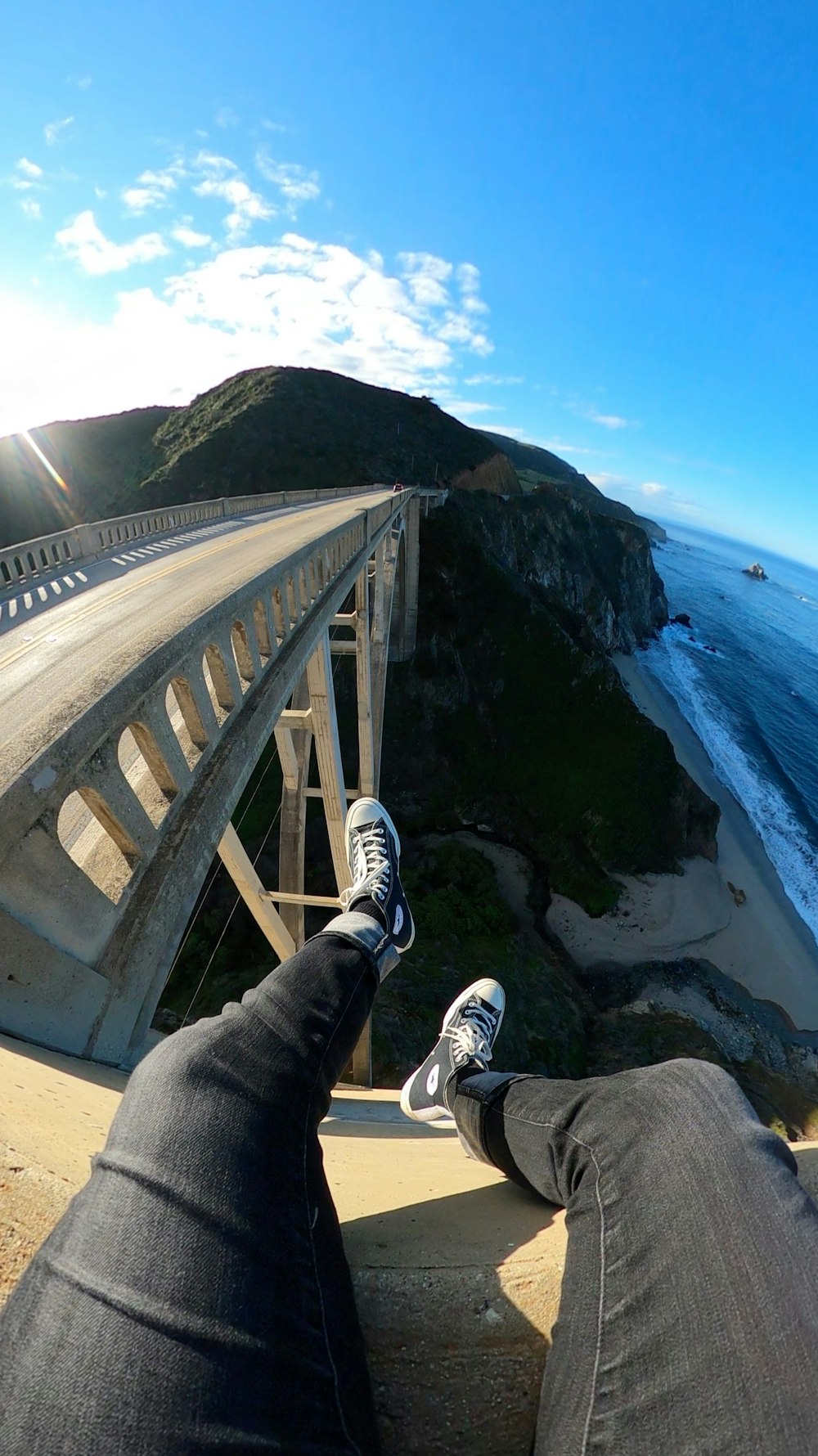 person in black pants and white sneakers sitting on rock near body of water during daytime