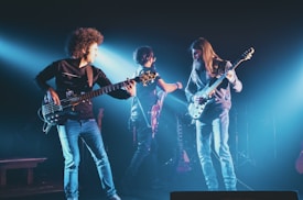 Three musicians are on stage playing electric guitars. They are surrounded by atmospheric blue lighting. The scene has a lively and dynamic energy, with the musicians focused on their performance.