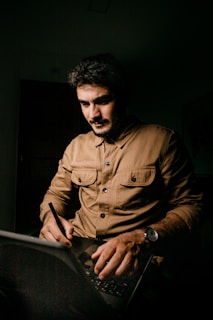 A person is sitting in a dimly lit room, focused on using a laptop and writing with a stylus. The individual is wearing a brown shirt and a watch, with dark hair and a beard. The light casts dramatic shadows, emphasizing concentration.