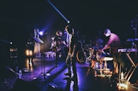 A band is performing on a dimly lit stage, with members playing various instruments such as guitar, keyboard, and drums. Stage lights on stands create dramatic shadows and highlight some band members against the dark background.