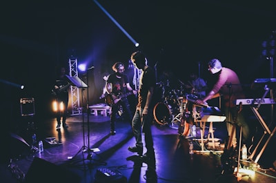 A band is performing on a dimly lit stage, with members playing various instruments such as guitar, keyboard, and drums. Stage lights on stands create dramatic shadows and highlight some band members against the dark background.