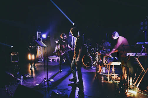 The band jamming together in a dimly lit rehearsal space, shadows playing across their instruments.