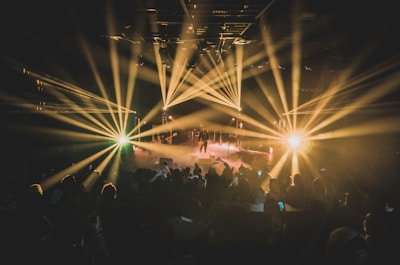 Colorful stage lights create dramatic beams across a crowded concert venue. The performance space is filled with fog, enhancing the vibrant effect of the lights. The audience is dimly lit, focused on the stage where musicians perform.