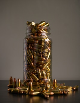 A glass jar filled with brass and copper-colored bullets, overflowing onto a dark surface. The background is neutral, highlighting the shiny metallic appearance of the ammunition.