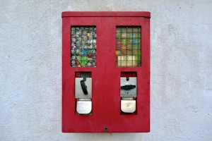 A red vending machine with two slots, featuring a grid-like display area showing various colorful items and numbers. The machine is mounted on a plain, light-colored wall.