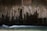 Dark, textured concrete wall with streaks and stains from water erosion, creating an abstract pattern. A stream of water flows smoothly at the bottom with a long exposure effect, creating a soft, blurred motion.