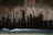 Dark, textured concrete wall with streaks and stains from water erosion, creating an abstract pattern. A stream of water flows smoothly at the bottom with a long exposure effect, creating a soft, blurred motion.