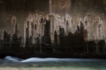 Dark, textured concrete wall with streaks and stains from water erosion, creating an abstract pattern. A stream of water flows smoothly at the bottom with a long exposure effect, creating a soft, blurred motion.