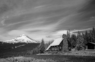 A rustic wooden cabin with a steep roof sits near a dense forest of tall coniferous trees. In the background, there is an impressive snow-capped mountain under a sky filled with wispy clouds.