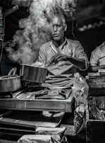 A man is cooking on a portable stove with a lot of steam rising from the pot, partially obscuring his face. The setting appears to be a street food stall or a small kitchen counter, with various packaged snacks and goods around. The lighting is dim, giving a moody atmosphere.