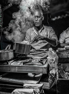 A man is cooking on a portable stove with a lot of steam rising from the pot, partially obscuring his face. The setting appears to be a street food stall or a small kitchen counter, with various packaged snacks and goods around. The lighting is dim, giving a moody atmosphere.