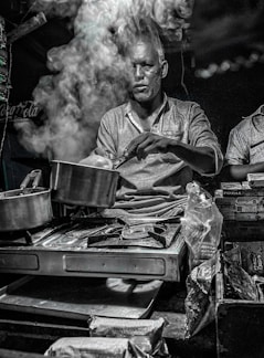 A man is cooking on a portable stove with a lot of steam rising from the pot, partially obscuring his face. The setting appears to be a street food stall or a small kitchen counter, with various packaged snacks and goods around. The lighting is dim, giving a moody atmosphere.
