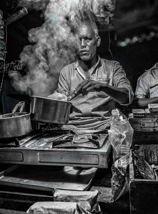 A man is cooking on a portable stove with a lot of steam rising from the pot, partially obscuring his face. The setting appears to be a street food stall or a small kitchen counter, with various packaged snacks and goods around. The lighting is dim, giving a moody atmosphere.