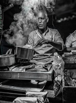 A man is cooking on a portable stove with a lot of steam rising from the pot, partially obscuring his face. The setting appears to be a street food stall or a small kitchen counter, with various packaged snacks and goods around. The lighting is dim, giving a moody atmosphere.