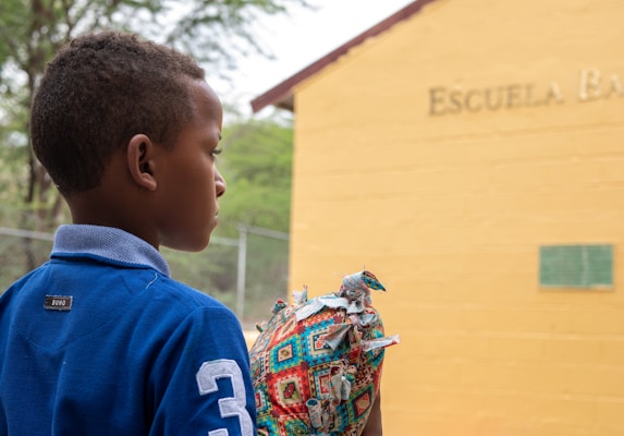 A young boy is standing outdoors facing a yellow building with the word 'Escuela' partially visible. He is wearing a blue shirt with a number on the sleeve and is holding a colorful, patterned object. The background includes a chain-link fence and some greenery.