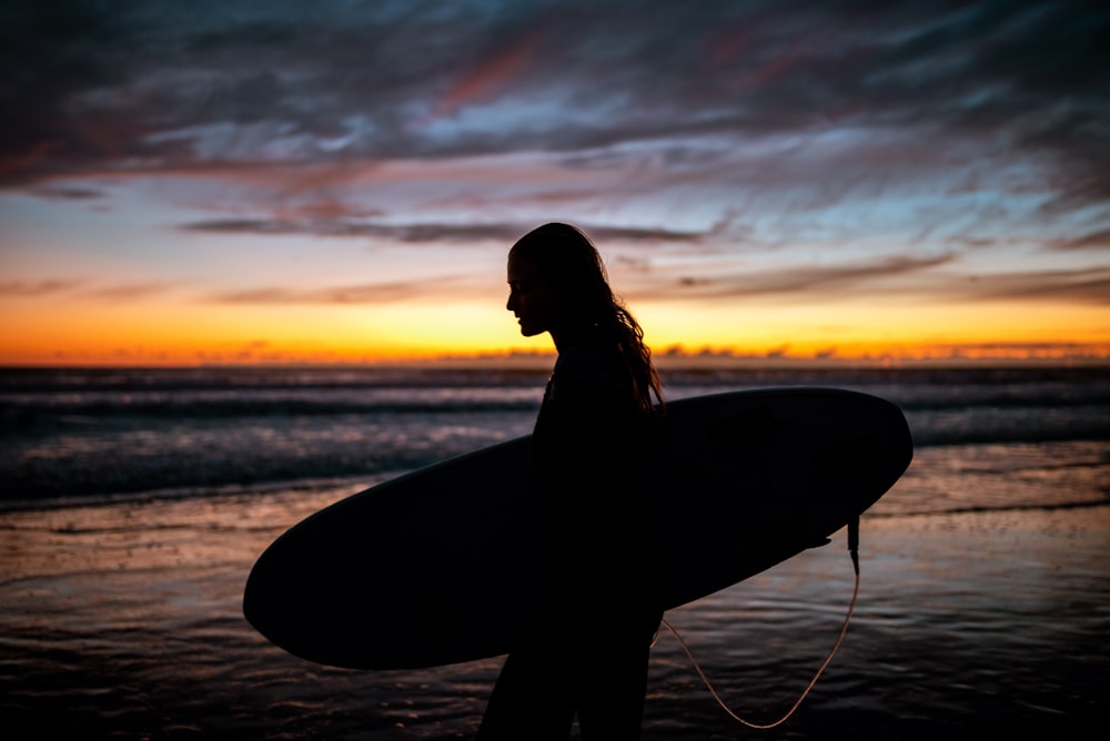 silhouette of woman holding surfboard during sunset