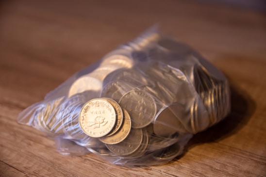 A plastic bag filled with coins rests on a wooden surface, with several coins prominently displaying engravings and designs, including currency markings and text.