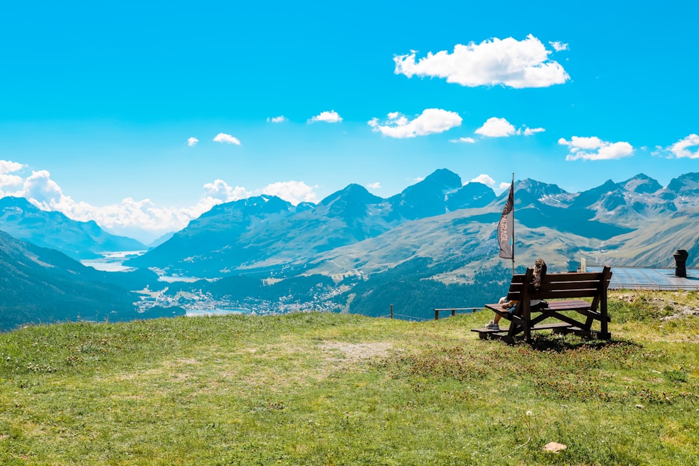 昼間は青空の下、山の近くの緑の芝生に茶色の木製のベンチ