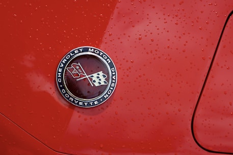 A close-up view of an emblem with the text 'Chevrolet Motor Division Corvette' featuring two crossed flags on a glossy red car surface with water droplets scattered across.
