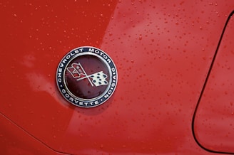 A close-up view of an emblem with the text 'Chevrolet Motor Division Corvette' featuring two crossed flags on a glossy red car surface with water droplets scattered across.