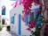 A charming white building with bright blue shutters and doors, adorned with vibrant pink bougainvillea climbing the walls. A small table and chairs sit under a shaded area, and the scene captures a sunny, Mediterranean atmosphere.