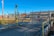 A secured gate covered with barbed wire, marked as Gate F, blocks a road leading to an industrial facility. Various warning signs are displayed, including a 'No Trespassing' notice. The background features industrial buildings and clear blue skies.