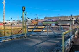 A secured gate covered with barbed wire, marked as Gate F, blocks a road leading to an industrial facility. Various warning signs are displayed, including a 'No Trespassing' notice. The background features industrial buildings and clear blue skies.